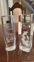 Two hexagonal clear glass pillars with laser-etched animal images inside, placed on a wooden table with a chair and staircase visible in the background.