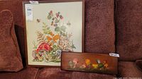 Framed mushroom needlework tapestry and wood board mushroom painting on couch showing size relative to couch cushions.