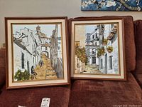 Two framed paintings side by side on upholstered surface, both architectural street scenes