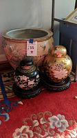 Photo showing two ginger jars (one black with floral and butterfly motifs, one gold with floral designs) and large round planter with koi fish and lotus flower decorations on patterned rug