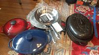 Set displayed on floor showing red and blue enameled cast iron pots with lids, stainless steel strainer, and other pans including covered black roasting pan.