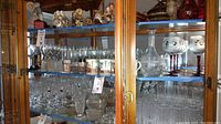 View of glassware collection inside display cabinet including clear crystal glasses, Irish Duiske glasses, decorative stemware, Lenox Disney mugs, and glass decanter.