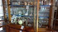 Full view of wooden display cabinet showcasing various glassware, plates, and porcelain figurines inside.