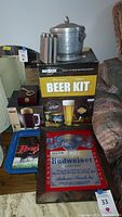 Wide view of items showing Budweiser serving tray, beer kit box, aluminum ice bucket with lid, and stainless steel flask on top. Mirror and other items visible in background.