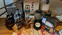 Photo showing multiple knife blocks with knives, Breville juicer box, blender with glass container, and West Bend cooker box against carpet background.