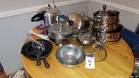 Wide view of the lot showing pressure cooker, stacked pots, cast iron pans, wire strainers, cutting boards, and utensils on a wood table.