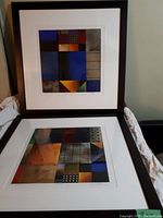 Photo of two framed abstract artworks stacked vertically, showing geometric shapes and muted colors with white matting and dark frames