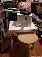 Photo showing the white plastic folding step stool placed atop the white TV tray table, with the round wooden stool nearby on wooden flooring.