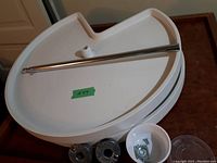 Two kidney-shaped white plastic corner shelves stacked, with a central hole for rod installation. A chrome metal support rod is placed on top. Hardware includes plastic spacers and metal fasteners in a small bowl for mounting.