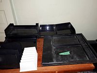 Photo shows multiple black plastic desk organizers arranged on a wooden surface including stackable flat paper trays, large multi-section flat organizer, and drawer organizer.
