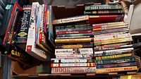 Photo showing a large collection of English children and youth books stacked inside two cardboard boxes on a wooden surface. Titles and authors visible include 'Oxford English Dictionary For Schools,' 'My Parents are Vampires,' 'Treasure Hunters,' and 'Bran Hambric'.