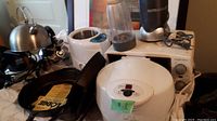 Photo showing 3 kettles including an electric wired stainless steel kettle, 2 new iron fry pans (one flat, one ridged), used NutriBullet blender, used white desktop oven, and white electric fry pan.