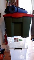 Stacked three plastic storage bins, large green bin on top with red lid, two clear bins below, two rolled yoga mats placed on top of the green bin