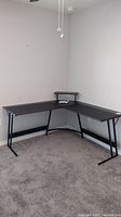 Full view of the black L-shaped desk positioned in the corner of the room, showing metal legs and elevated monitor stand.