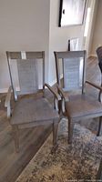 Pair of two wooden dining chairs with armrests and brown upholstered seats in room with hardwood floor and patterned area rug.