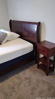 Full view of dark wood queen sleigh bed headboard and side cabinet with drawer and two shelves placed on beige carpet. Includes pillows and partial view of mattress.