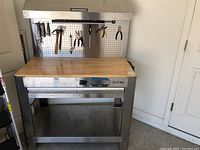 Front view of the stainless steel work bench with wooden top, two drawers, bottom storage shelf, and pegboard with hanging tools.