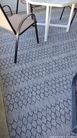 Photo of outdoor area rug partially under white metal table and black metal chairs, showing geometric hexagonal-like black design on gray background.