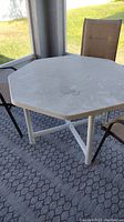View of the entire octagonal white plastic table on outdoor rug with some chairs visible in the background. Tabletop surface shows marks and dirt suggesting it needs cleaning.