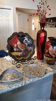 Photo showing all items in the lot together on a countertop with a white background wall, including two red and black vases with rooster designs, the rooster platter on metal easel, and the two handmade pottery dishes with swirl patterns.