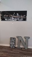Photo showing full view of the wall art items together: New York skyline canvas print, metal letter N, and inspirational sign on floor against white wall.