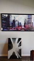 Large horizontal city skyline canvas wall art with reds, blues, and purples mounted on the wall above a smaller black and white vertical city skyline canvas leaning against the wall.