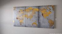 Three-piece canvas wall art showing world map in gold leaf style on gray textured background, hung on white wall.