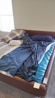 Full view of king size waterbed with blue water mattress visible under dark blue bedding and multiple pillows. Brown faux leather headboard and side rails visible.