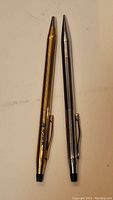 Two ballpoint pens, one gold-colored and one silver-colored, shown tip up to display the clip and button mechanisms.