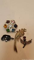 Four costume jewelry brooches: multicolor gem cluster flower, black fan-shaped with rhinestones, gold-tone floral branch, and black/silver thistle with purple rhinestone.