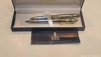 Set of Bill Blass pen and pencil lying inside open black presentation box with light lining and white ribbon strap, with a Bill Blass information card.