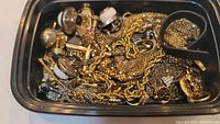 Overview of mixed costume jewelry in plastic container including chains, earrings, rings.
