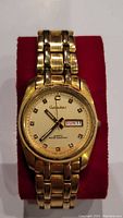Front view of gold tone Garsedina wristwatch on red cushion, showing cream dial with day-date indicator.