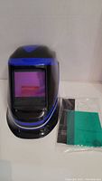 Front view of the DEKO welding helmet showing protective viewing window and accessories (instructions, battery).