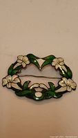 Front view of oval sterling silver brooch with white flower and green leaf enamel decoration on white background.