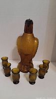 Frontal view of the eagle-shaped decanter centered with six matching amber shot glasses arranged around it.