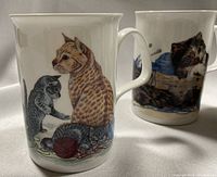 Close-up showing multiple mugs with detailed cat illustrations including cats playing with yarn and resting.