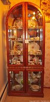 Front view of curio display cabinet filled with porcelain dishware and teacups. Wooden frame with glass doors and mirror back visible.
