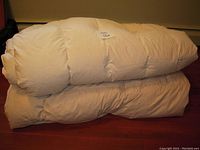 Rolled and stacked view of two off-white down comforters on a wooden floor.