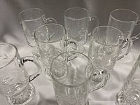 Eight clear glass mugs with pedestal bases showing etched leaf design, grouped together on white fabric backdrop.
