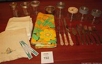 Full lot showing vintage tablecloths, fabric napkins, collectible jars, and assorted cutlery arranged in rows on a wooden table.