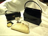 Three vintage purses: a large black patent leather handbag needing repair, a smaller black leather handbag with a gold twist-lock clasp, and a gold metallic evening clutch with a chain strap, displayed on cream fabric.