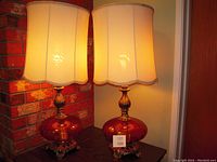 Two amber glass base table lamps with ornate metal feet and fabric shades, placed on wooden table by brick wall.
