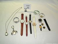 Photo shows entire assortment of 13 watches and watch accessories displayed on white surface, including pendant watch, ladybug keychain watch, wristwatches with leather and plastic bands, and loose watch faces and bands.