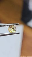 Close-up of one round cut yellow-green sphene gemstone held by tweezers showing its adamantine luster and brilliance.