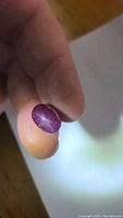 Close-up photo of deep purple-red oval cabochon natural star ruby being held between fingers showing surface inclusions and star effect under light.