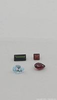 Four loose natural gemstones on a white background showing a light blue aquamarine, dark green tourmaline, reddish garnet, and dark red garnet.
