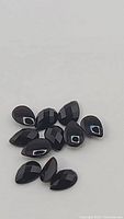 Close-up of 11 pear shaped black onyx checker cut gemstones showing facets and size relative to camera.