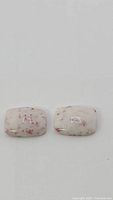 Pair of natural Mexican cinnabar cushion cabochon gemstones shown on light background with close up detail visible. Each stone has a white/cream colored base with red cinnabar speckles.
