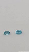 Close-up photo of two oval-cut blue apatite gemstones on a white background, showing fine facets and fluorescent Paraiba blue color.